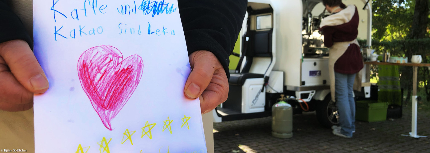 Ein selbstgemaltes Kinderbild mit Herzen wird von zwei Händen gehalten, im Hintergrund steht Kaffeemobil PAUL