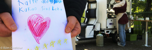 Ein selbstgemaltes Kinderbild mit Herzen wird von zwei Händen gehalten, im Hintergrund steht Kaffeemobil PAUL