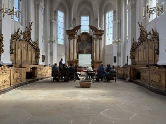 Ein Alttarraum mit hohen Kirchenfenstern, davor sitzen Menschen in Stuhlreihen für einen Workshop