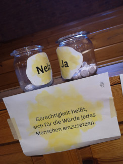 Zwei Gläser mit Aufdruck "nein" und "ja", in dem Glas mit "ja" sind viele Steine, in dem mit "nein" wenige Steine. Auf einem Schild steht: "Gerechtigkeit heißt, sich für die Würde jedes Menschen einzusetzen