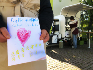 Ein selbstgemaltes Kinderbild mit Herzen wird von zwei Händen gehalten, im Hintergrund steht Kaffeemobil PAUL