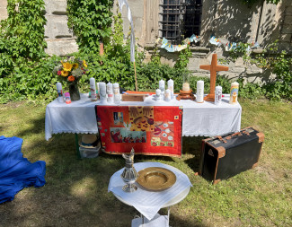 ein Altar im Grünen, geschmückt mit zahlreichen Taufkerzen; ein altertümlicher Koffer steht im Vordergrund, auch eine Taufschale
